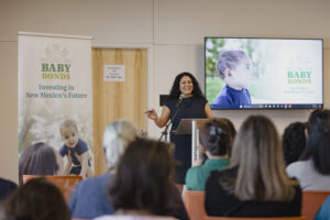 New Mexico State Treasurer Laura M. Montoya presents the Baby Bonds Program. She is smiling in a blue dress behind a podium. She is gesturing towards a large poster and projector displaying images of happy children and happy parents. The audience is intrigued and focusing with full attention at the presentation from the State Treasurer.